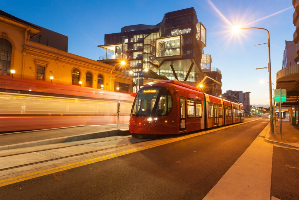 A Newcastle light rail tram at dusk on Hunter Street, with the University of Newcastle NeW Space building lit up behind it.